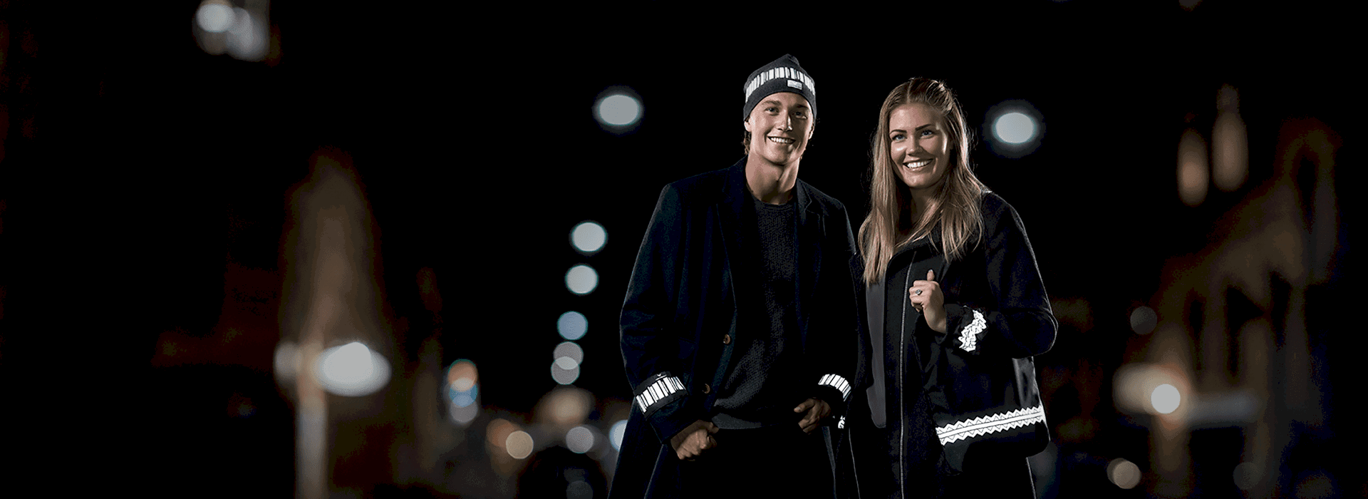 Man och kvinna. Han bär mössa med reflexmönstret norrsken. Hon håller i Reflexväska med mönstret Fjälltopp. Båda bär ärmreflex